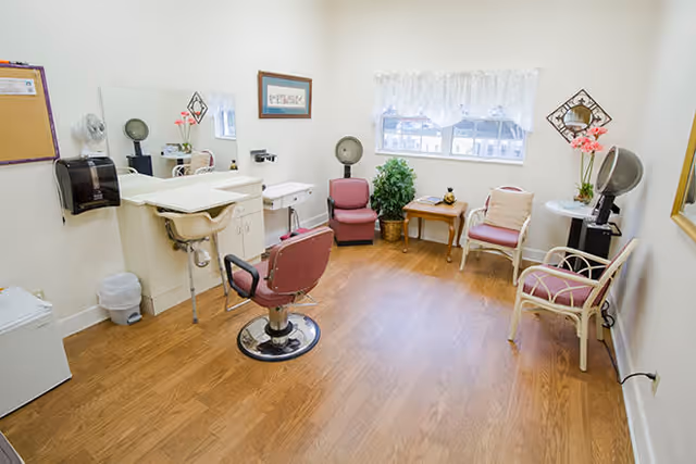 A bright room with wooden flooring featuring a salon chair in front of a sink and mirror setup. The room also has several chairs arranged around a small table near a window with sheer white curtains. There is a hair dryer on a stand, a potted plant, and a few decorative items including flowers and framed pictures on the walls.