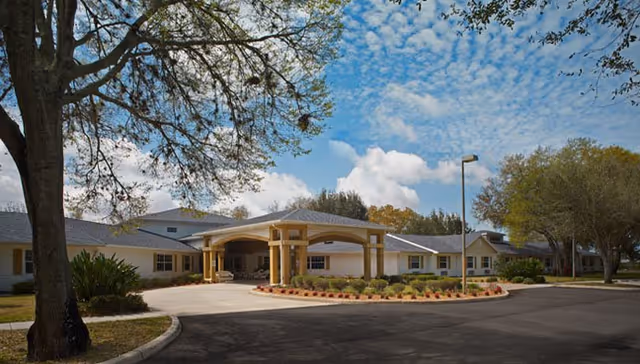 Exterior view of a single-story senior living facility with a covered entrance, surrounded by trees and landscaping under a partly cloudy sky.