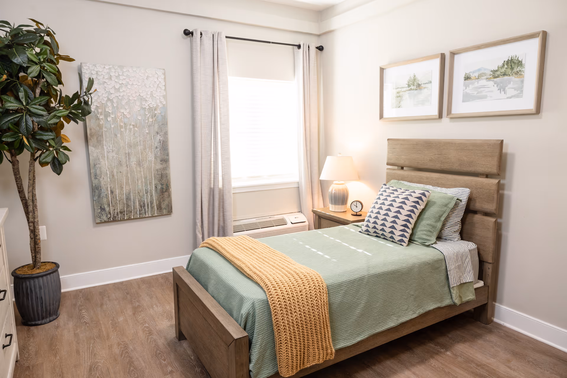 A neatly decorated single bedroom with a wooden bed, green bedding, a nightstand and lamp, wall art, and a potted plant.