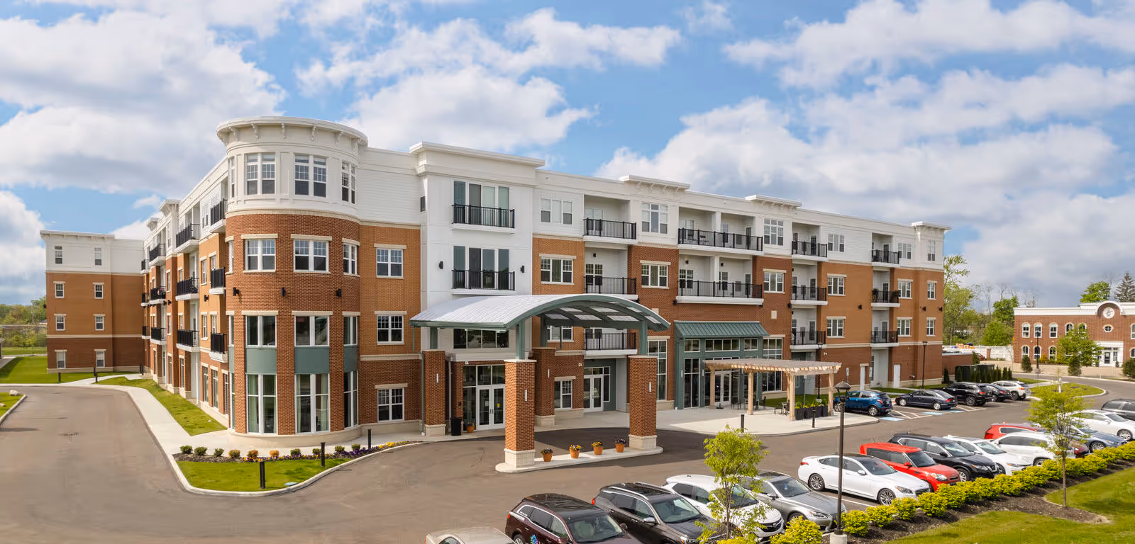 Four-story brick and white residential building with a covered main entrance and a parking lot with cars.