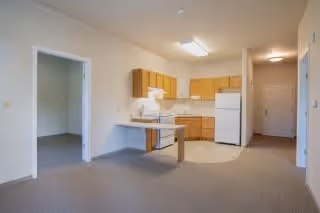 Interior view of a senior living apartment featuring a small kitchen area with wooden cabinets, a white refrigerator, stove, and microwave. The kitchen is adjacent to a carpeted living space with an open doorway leading to another room. The walls are painted light beige and the space is well-lit with ceiling lights.