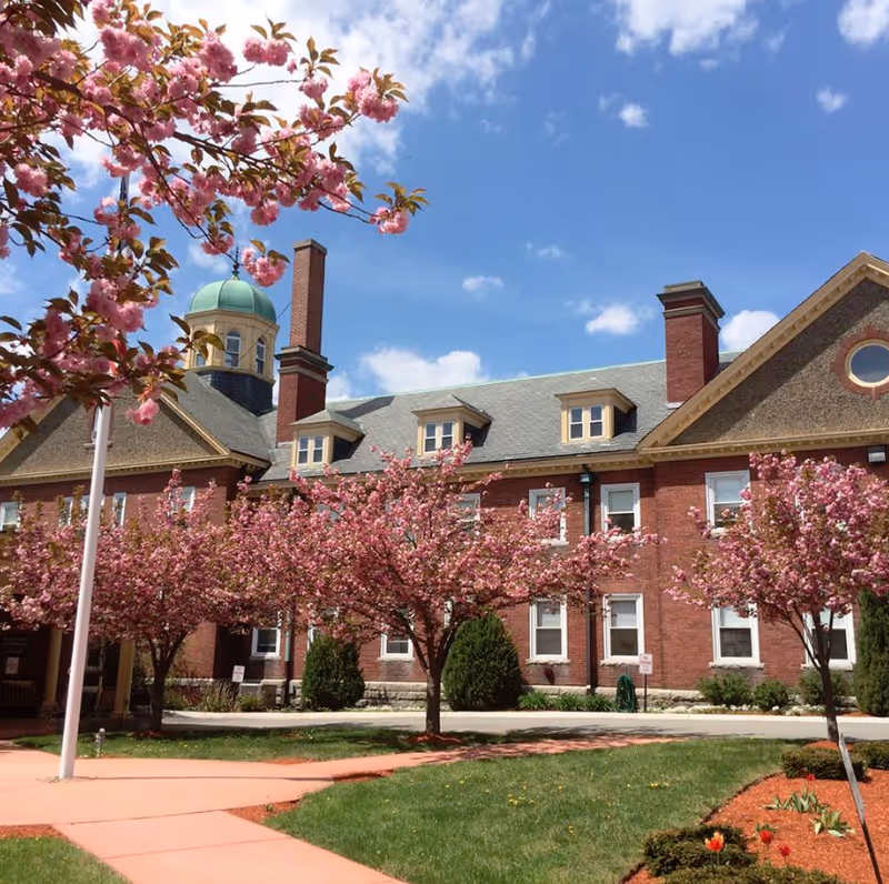 Exterior view of a red brick building with a green dome and multiple chimneys under a blue sky with scattered clouds. The foreground features a walkway surrounded by green grass and blooming pink cherry blossom trees.