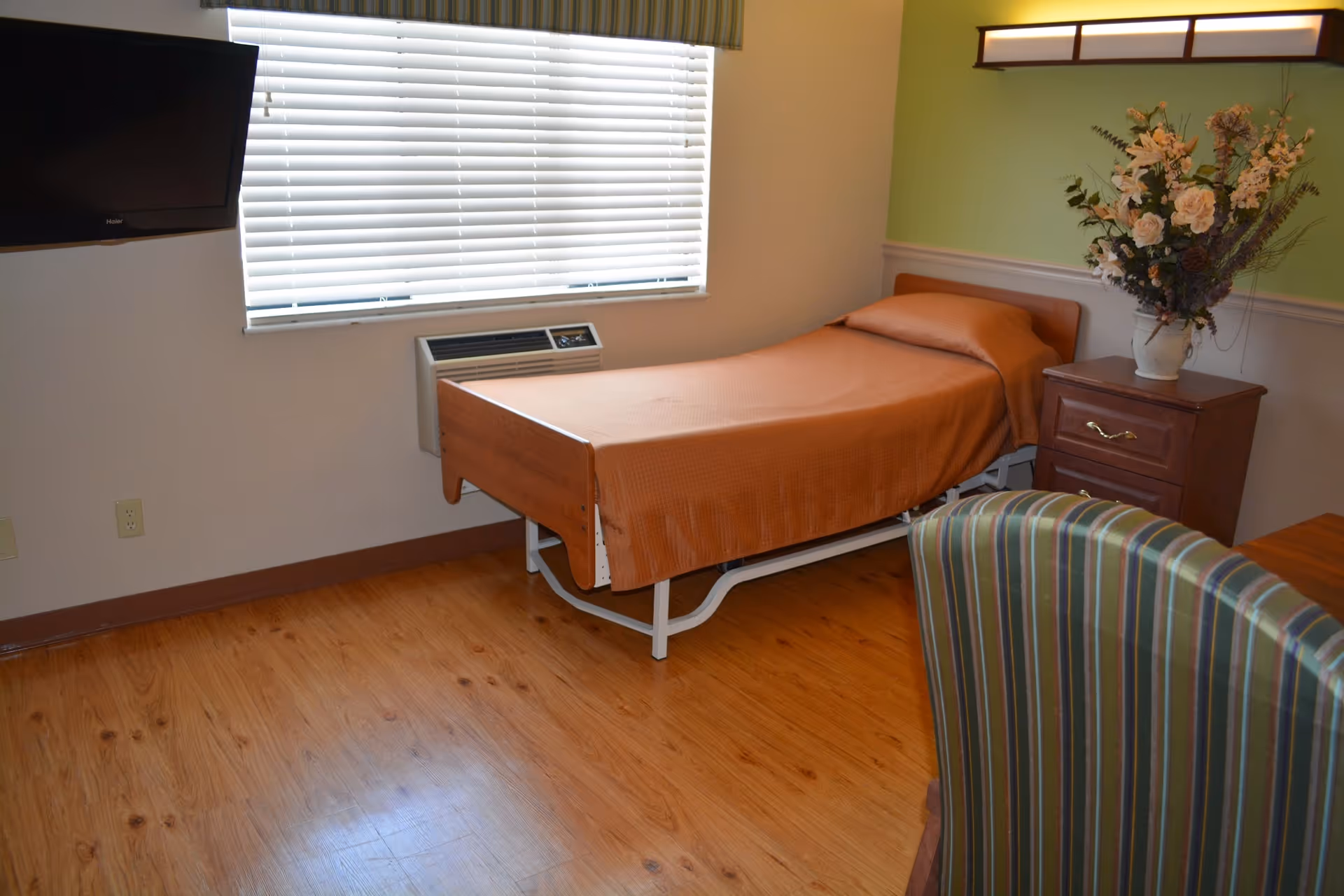 A simple nursing home bedroom with a single bed covered in a brown blanket, a wooden nightstand with a vase of flowers, a striped upholstered chair, a wall-mounted flat-screen TV, and a window with closed blinds.