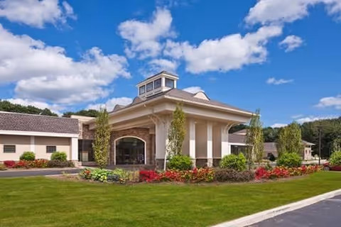 Exterior view of a single-story building with a covered entrance, surrounded by well-maintained landscaping including green grass, shrubs, and colorful flowers under a blue sky with scattered clouds.