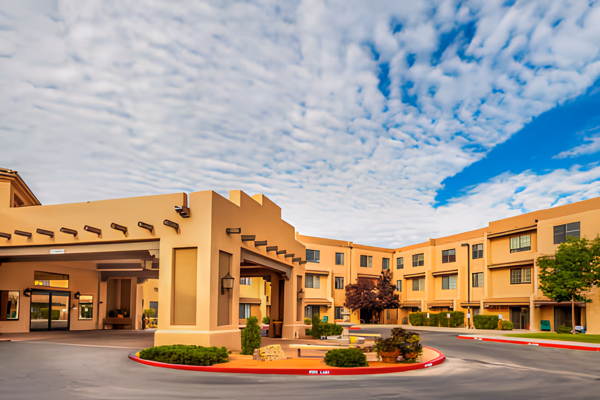 Exterior view of Fairwinds - Rio Rancho facility showing a tan-colored building with multiple windows, a covered entrance with wooden beams, landscaped bushes, and a partly cloudy blue sky overhead.