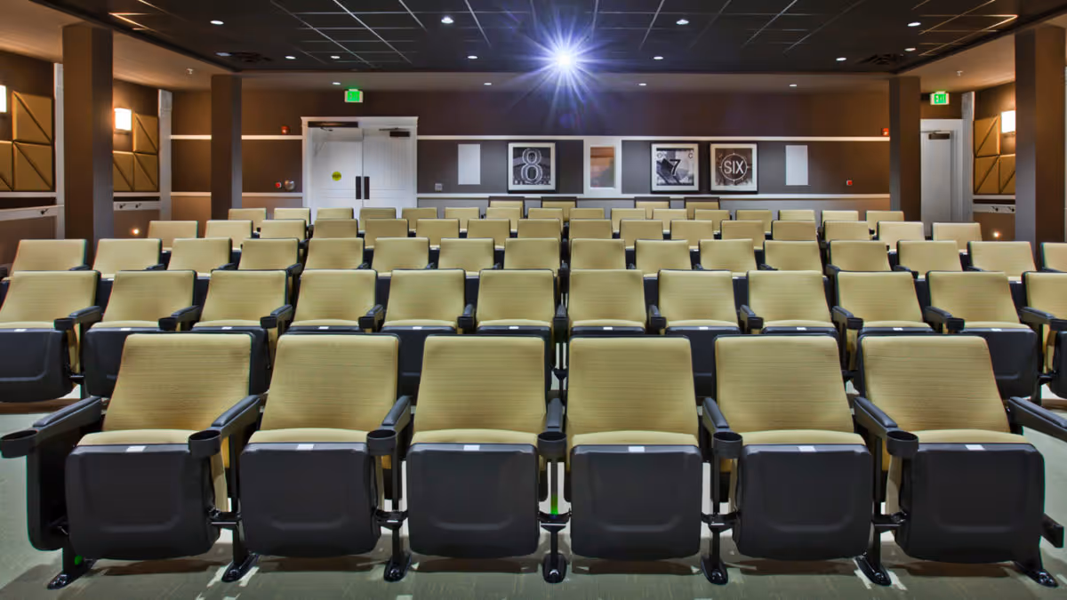 Rows of tan upholstered theater seats in a small auditorium facing a screen.