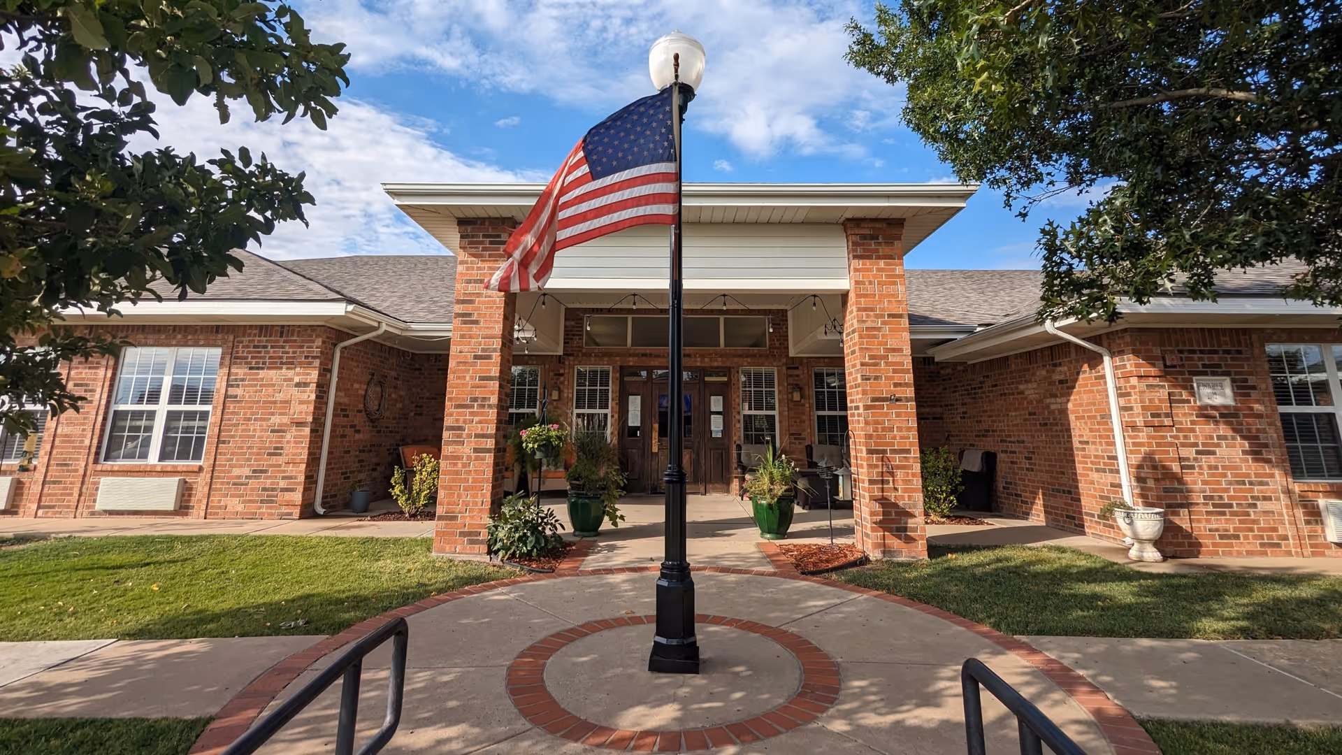 Front entrance of a brick building with a covered porch supported by two brick columns. An American flag is mounted on a black pole in the center of a circular brick and concrete walkway leading to the entrance. There are green plants in pots on the porch and trees framing the scene under a partly cloudy blue sky.