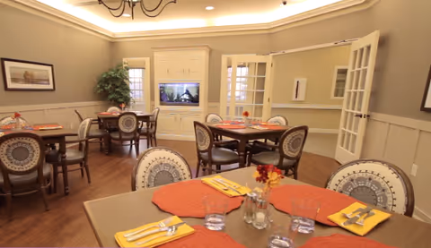 Interior view of the dining room at Colonial Oaks at Arlington, featuring wooden tables set with orange placemats, chairs with decorative backs, and a television in the background.
