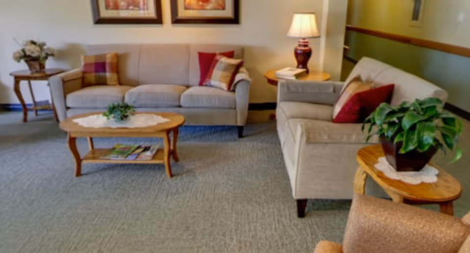 A cozy living room area with two beige sofas adorned with plaid and solid red throw pillows. A wooden coffee table with a lace doily and a small green plant sits in front of the sofas. There are two side tables, one with a lamp and books, and the other with a potted plant. The room has carpeted flooring and framed artwork on the wall.