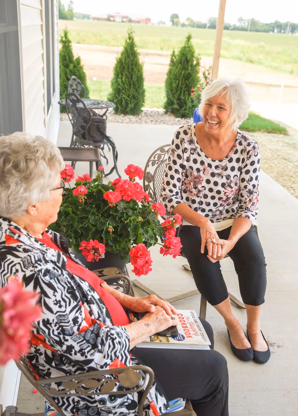 Two elderly women sitting and chatting on a covered outdoor patio with metal chairs and a table. One woman is holding a cookbook, and there are vibrant pink flowers in a pot between them. The background shows a grassy field and small evergreen shrubs.