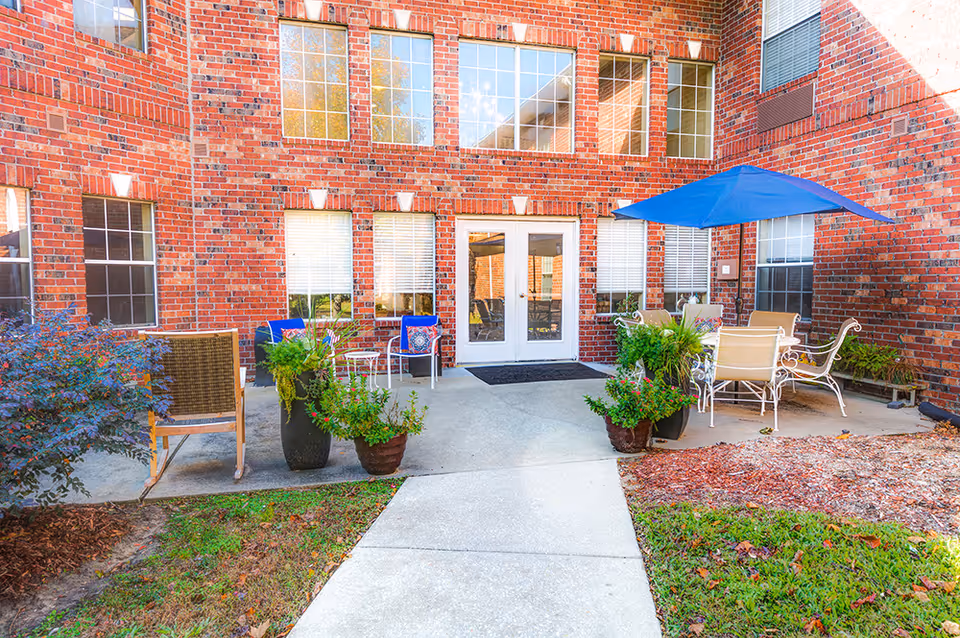 Outdoor patio area at Laketown Village with brick walls, multiple windows, a glass door, several potted plants, a blue patio umbrella shading a table with chairs, and additional seating including a rocking chair.
