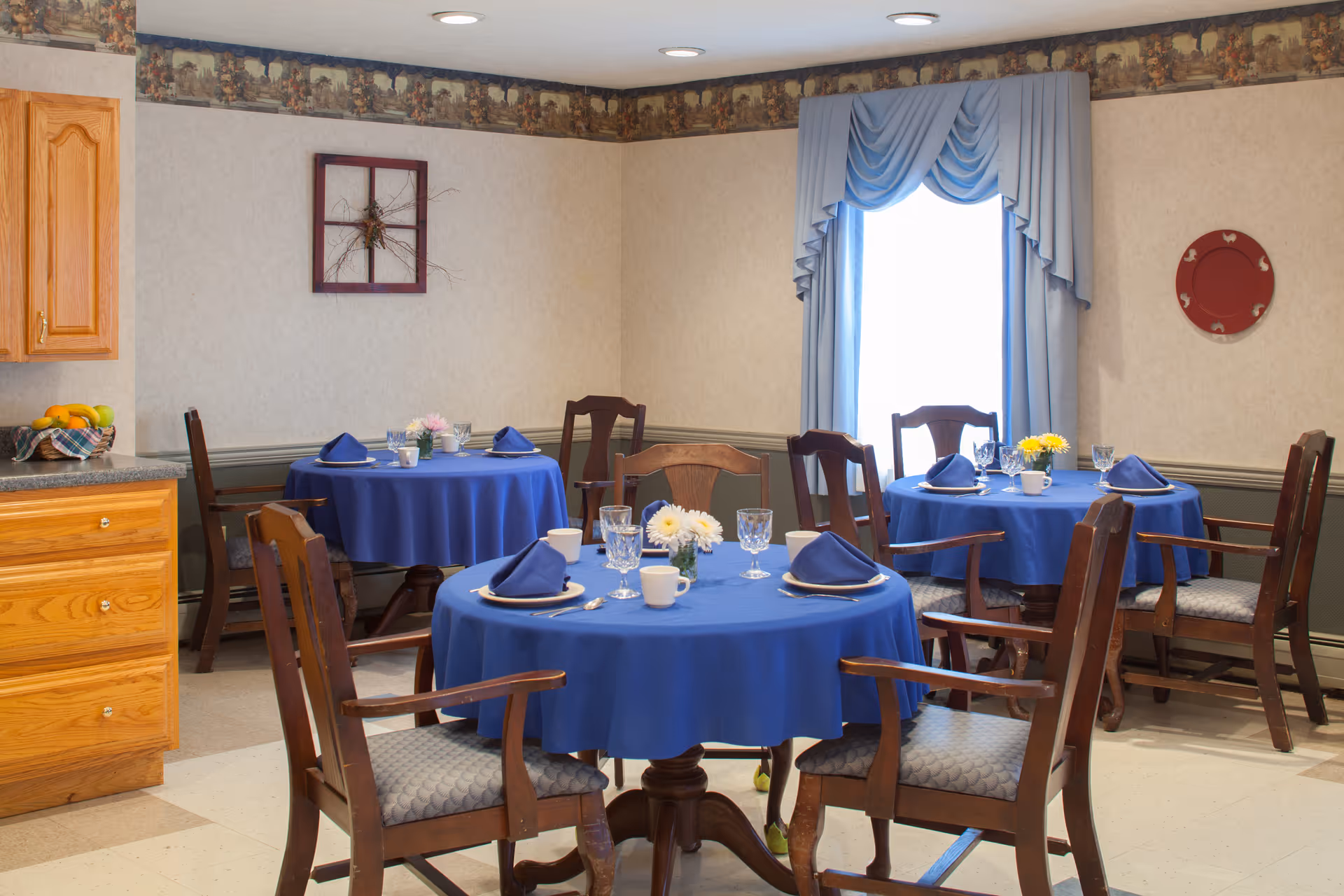 Small dining room with round tables set with blue tablecloths, napkins, glassware and wooden chairs near a window with blue drapes.