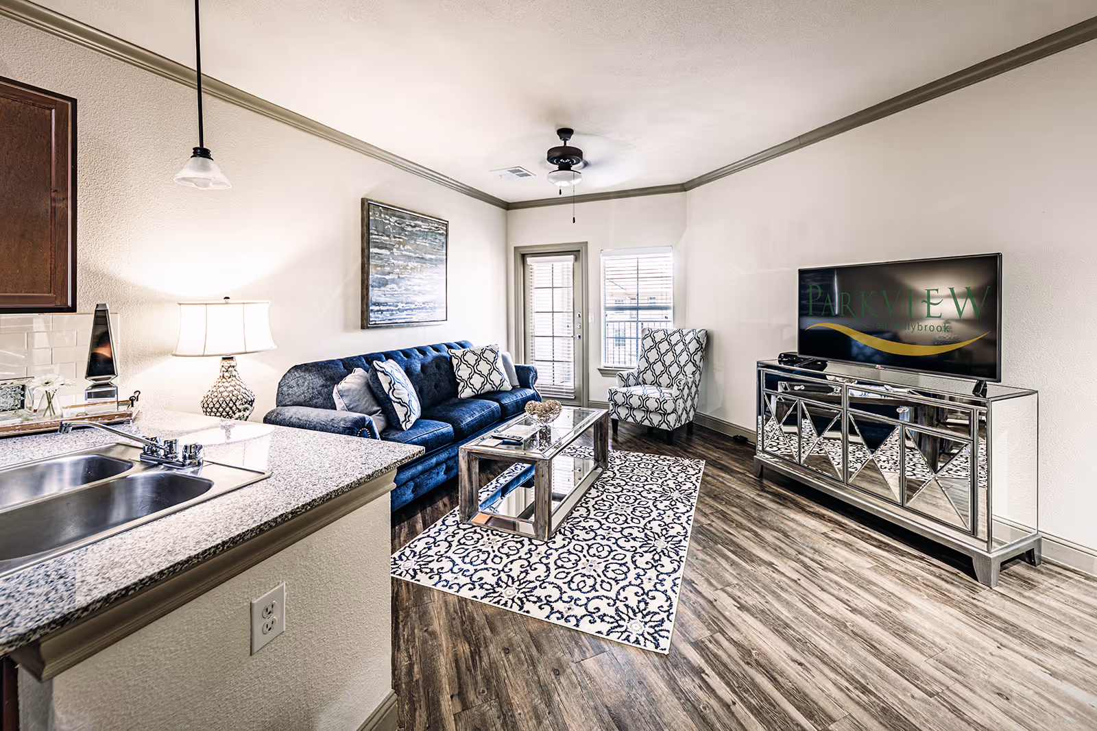 A modern living room with a blue sofa, patterned armchair, glass coffee table on a decorative rug, a mirrored TV stand with a flat-screen TV displaying the Parkview on Hollybrook logo, and a kitchen counter with a sink in the foreground.