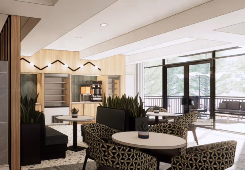 A bright and modern common area with round tables, patterned chairs, and black cushioned benches. There are potted plants on the tables and in planters between seating areas. The back wall features wooden cabinetry with a coffee machine and shelves. Large windows and glass doors open to an outdoor patio with additional seating and greenery visible outside.