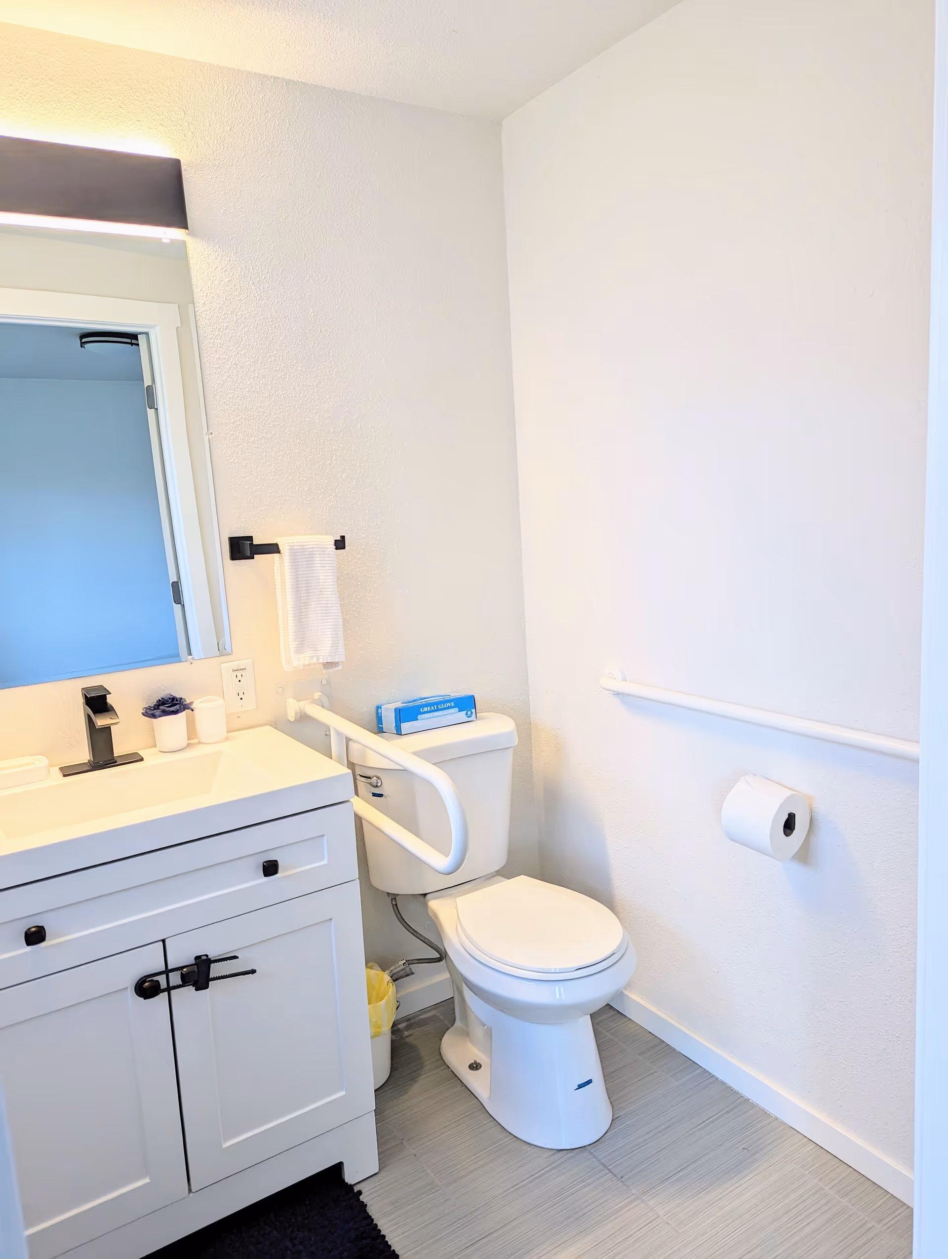 A clean and bright bathroom with a white toilet equipped with a safety grab bar on the left side. Next to the toilet is a white vanity with a sink, black faucet, and a mirror above it. A white towel hangs on a black towel holder mounted on the wall beside the mirror. The bathroom has light-colored tiled flooring and white walls, with a toilet paper holder mounted on the right wall.