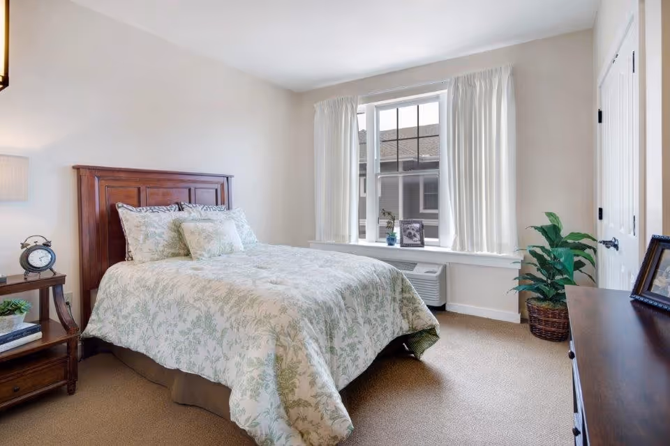A bright bedroom with a wooden headboard bed covered in a white and green floral patterned comforter and pillows. There is a wooden nightstand with a lamp, clock, and small plant on the left side of the bed. A large window with white curtains lets in natural light. A potted plant is placed near a white door on the right side, and a wooden dresser with framed pictures is partially visible in the foreground.