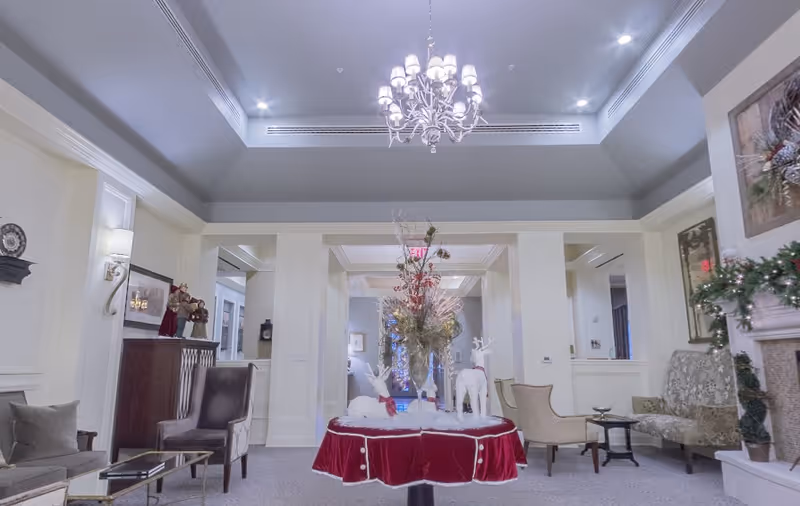 A spacious and elegantly decorated living room area with a chandelier hanging from the ceiling. In the center, there is a round table covered with a red velvet cloth and adorned with festive holiday decorations including reindeer figures and a floral arrangement. The room features comfortable seating including armchairs and a patterned sofa, with wall sconces and framed artwork on the walls. There is also a fireplace decorated with garland on the right side.