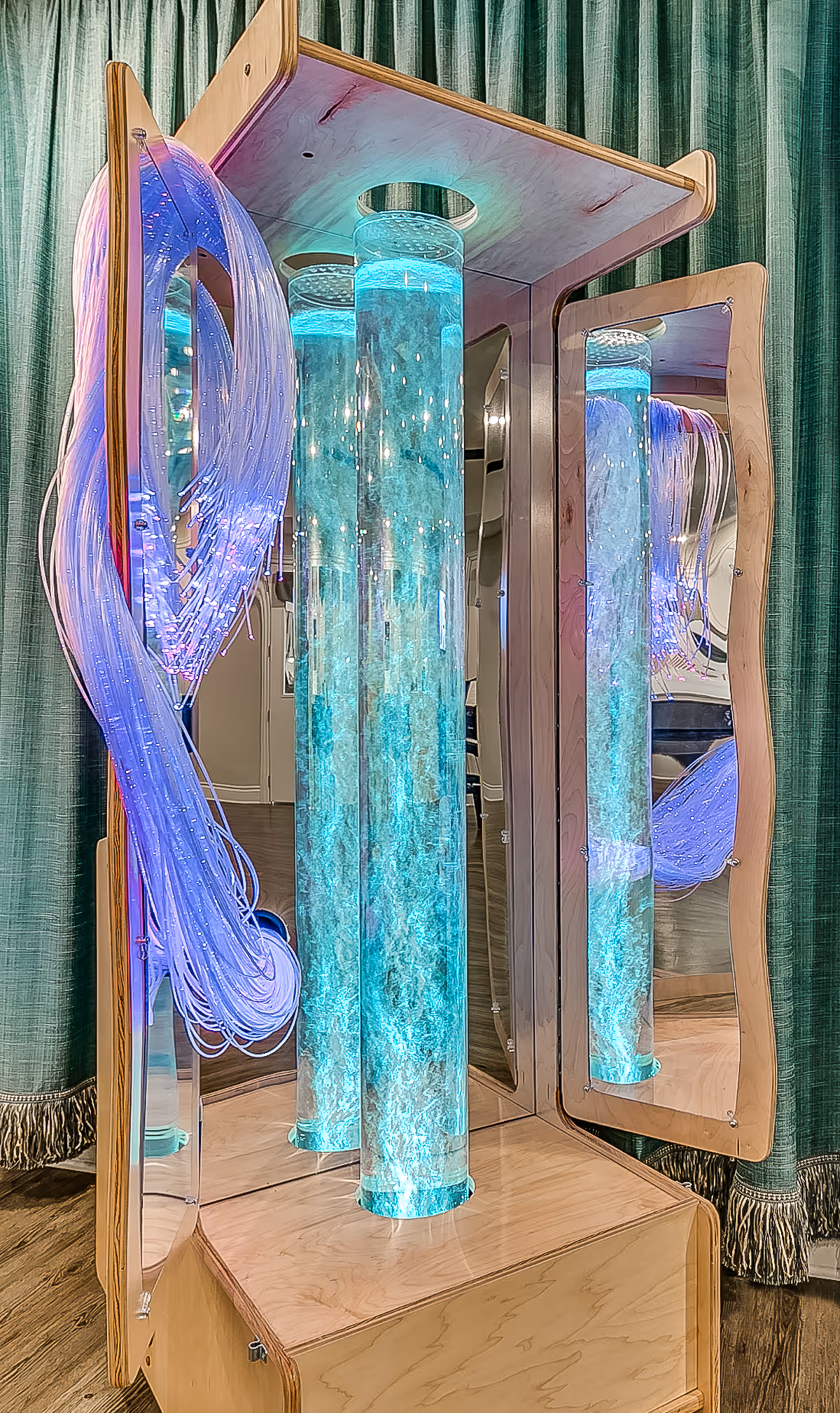 A sensory bubble tube with glowing blue water and bubbles inside, surrounded by fiber optic strands emitting purple light, mounted on a wooden base with mirrors on the sides reflecting the lights and colors, set against a green curtain background.