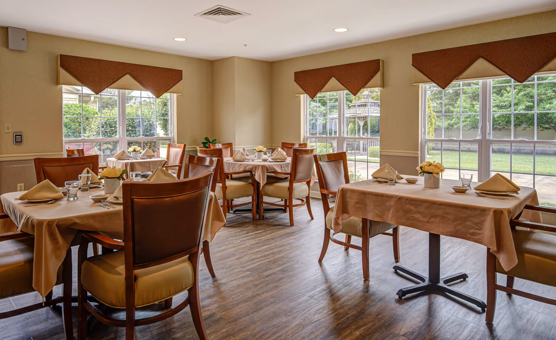 A bright dining room with several tables covered in beige tablecloths, each set with folded napkins, glasses, and small flower arrangements. The room has large windows with brown valances, allowing natural light to illuminate the wooden floor and chairs with cushioned seats.