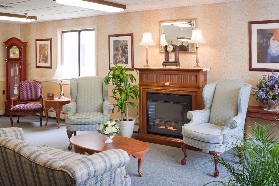 A cozy living room with a striped sofa, two light blue patterned armchairs, a wooden coffee table with a flower arrangement, a wooden fireplace with a clock and two lamps on top, a tall grandfather clock, framed paintings on the walls, and several green plants.