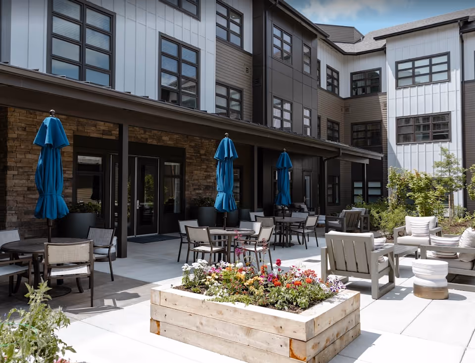 Outdoor courtyard with patio seating, tables, blue umbrellas, and a wooden planter in front of a multi-story senior living building.