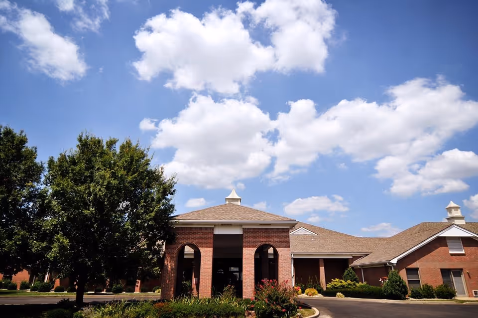 Exterior view of a single-story brick building with a covered entrance and a peaked roof, surrounded by green trees and shrubs under a blue sky with scattered white clouds.