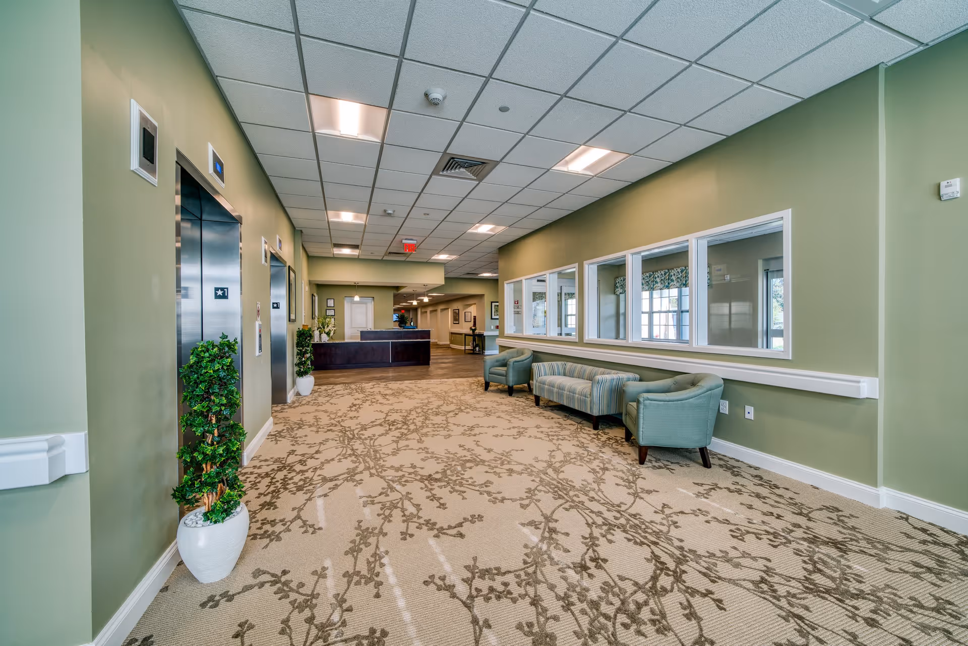 Spacious green-painted senior living facility interior hallway with seating, elevators on the left, and a reception desk ahead.