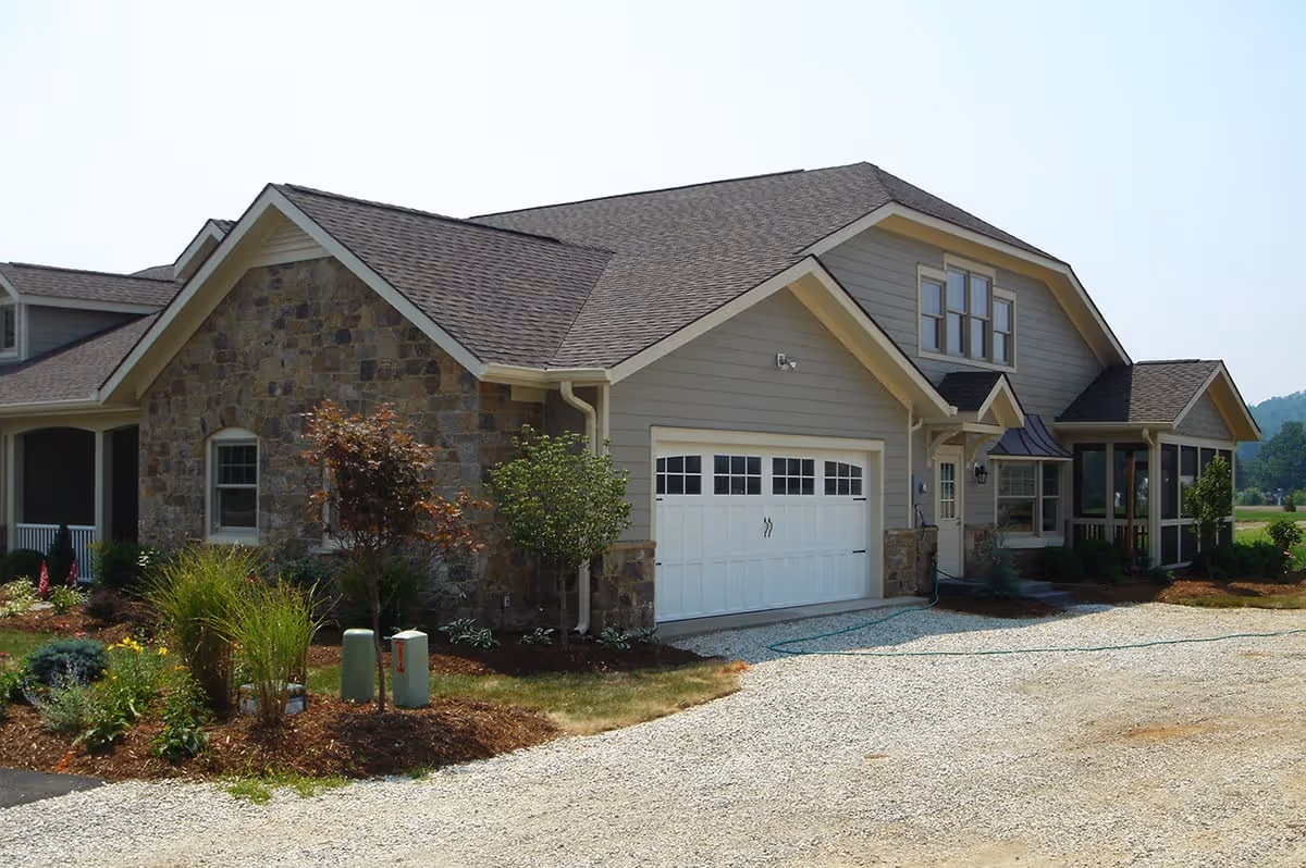 Exterior view of a single-story residential building with a stone and siding facade, a white garage door, and a gravel driveway. The building is surrounded by landscaped plants and small trees under a clear sky.