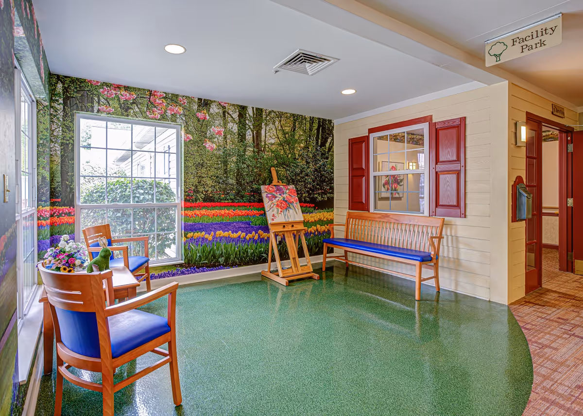 A bright and colorful indoor seating area with wooden chairs and benches featuring blue cushions. The walls are decorated with a large mural of a garden with flowers and trees, and there is an easel with a floral painting. A window with red shutters is part of the wall design, and a sign overhead reads 'Facility Park'.