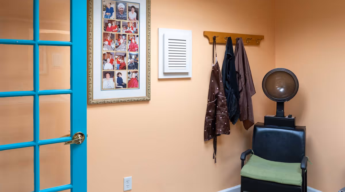A small room with peach-colored walls featuring a black salon chair with a green cushion and a hair dryer hood attached. On the wall above the chair, there is a wooden coat rack with three hanging aprons. To the left, a framed collage of photos of elderly people is displayed. A turquoise door with glass panes is partially visible on the left side of the image.