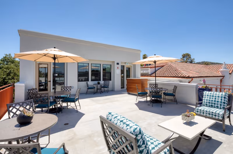 Rooftop patio with tables, chairs, umbrellas and lounge seating outside a light-colored building under a clear blue sky.