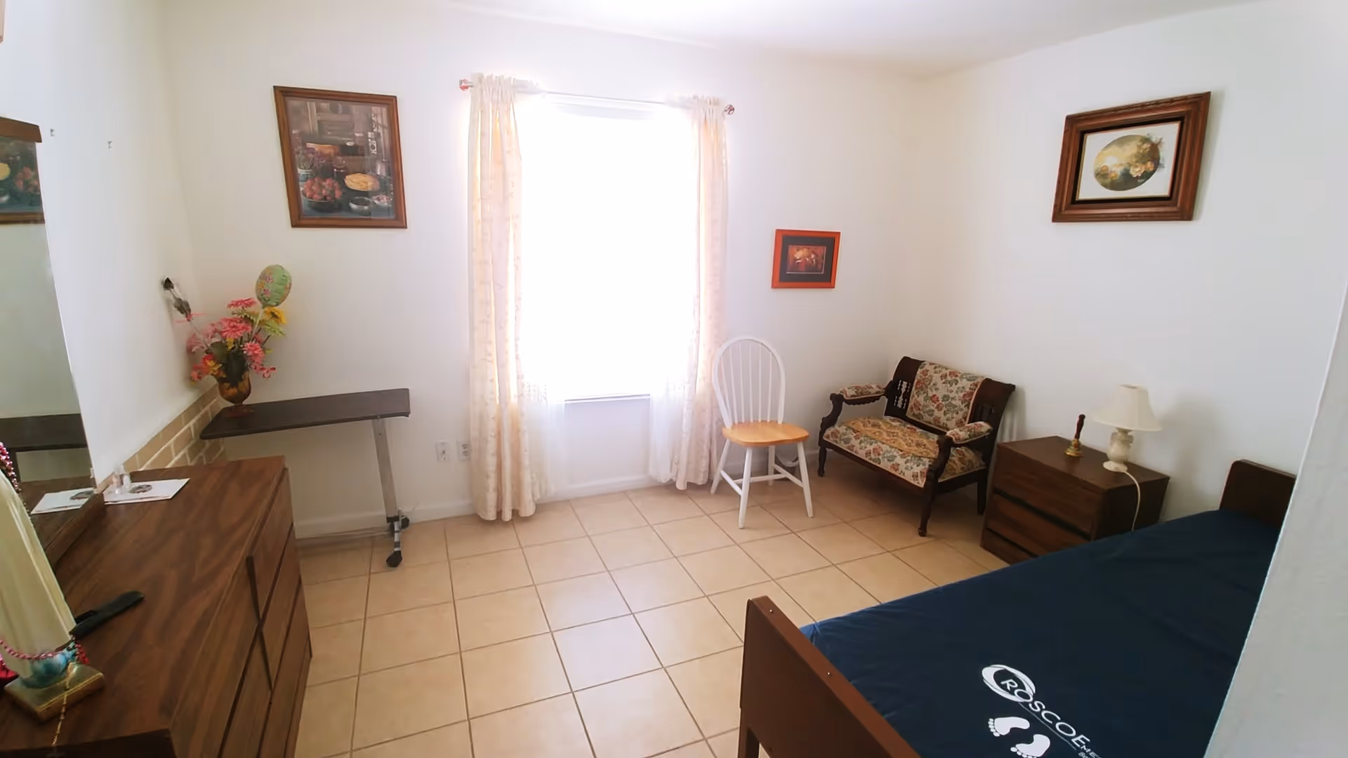 A simple bedroom with a single bed covered with a dark blue sheet, a wooden dresser with a mirror, a small wooden nightstand with a lamp, a floral upholstered loveseat, a white wooden chair, and a window with light curtains letting in natural light. The walls are white and decorated with framed pictures.