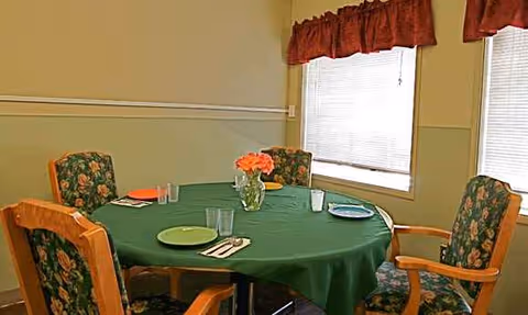 Round dining table with green tablecloth, a vase of orange flowers, and floral chairs.