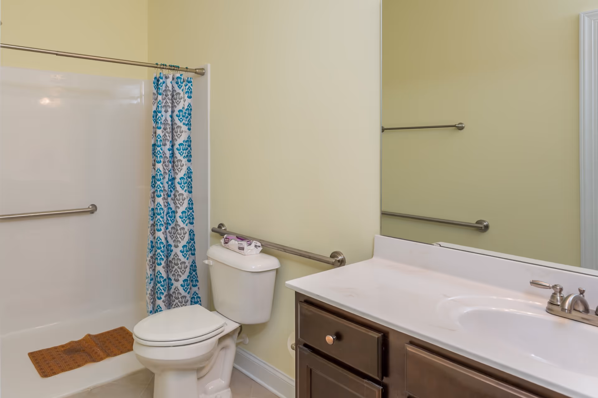 Bathroom with toilet, sink, shower with a blue patterned curtain, grab bars, and a large mirror.
