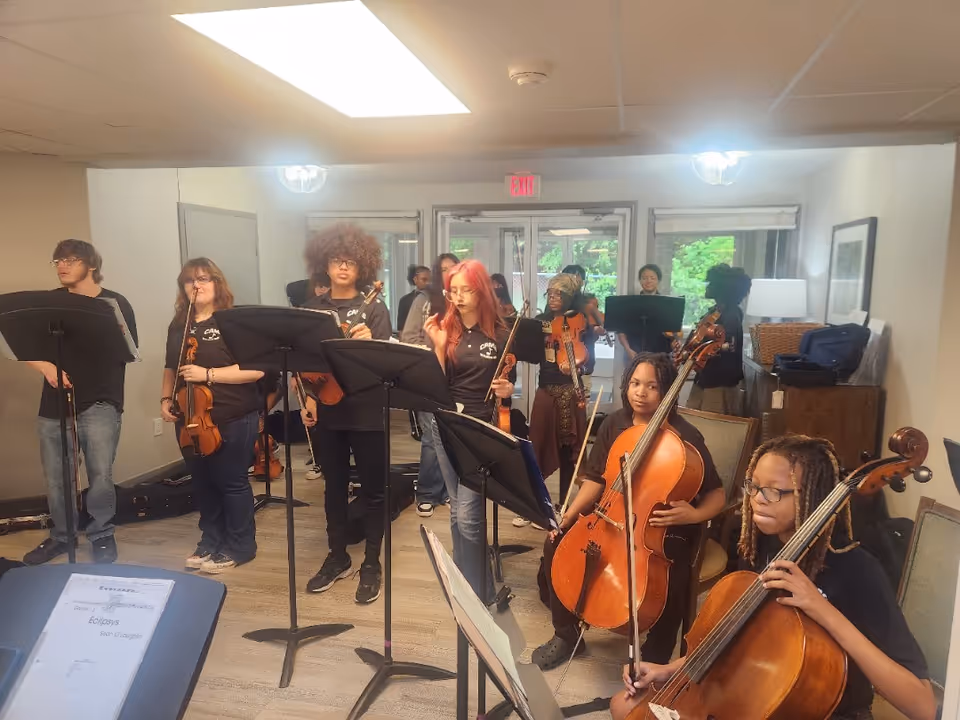A group of young musicians practicing indoors, some standing with violins and others seated with cellos, in a well-lit room with music stands and sheet music.