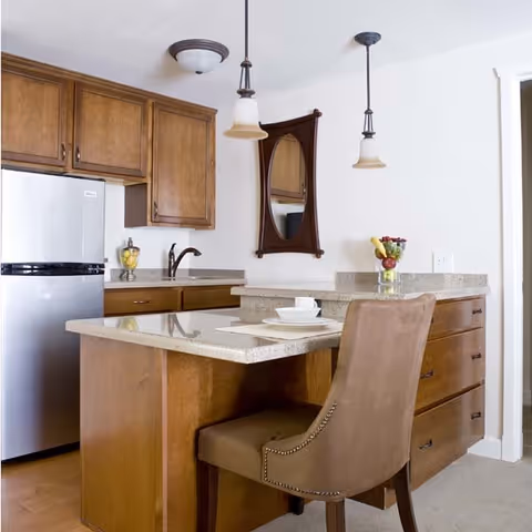 Modern kitchen with wooden cabinets, a stone countertop island with a padded chair, pendant lights, and a refrigerator.