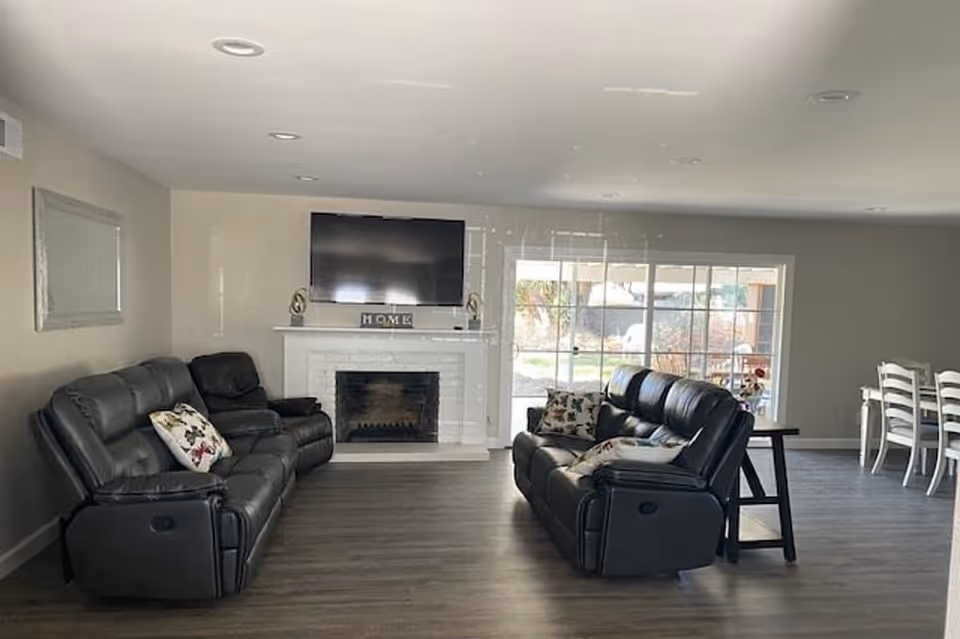 A spacious living room with two black leather sofas facing a white fireplace with a mounted flat-screen TV above it. The fireplace mantle has decorative items including a block sign spelling 'HOME'. There is a large window behind one sofa letting in natural light, and a dining area with white chairs is visible in the background.