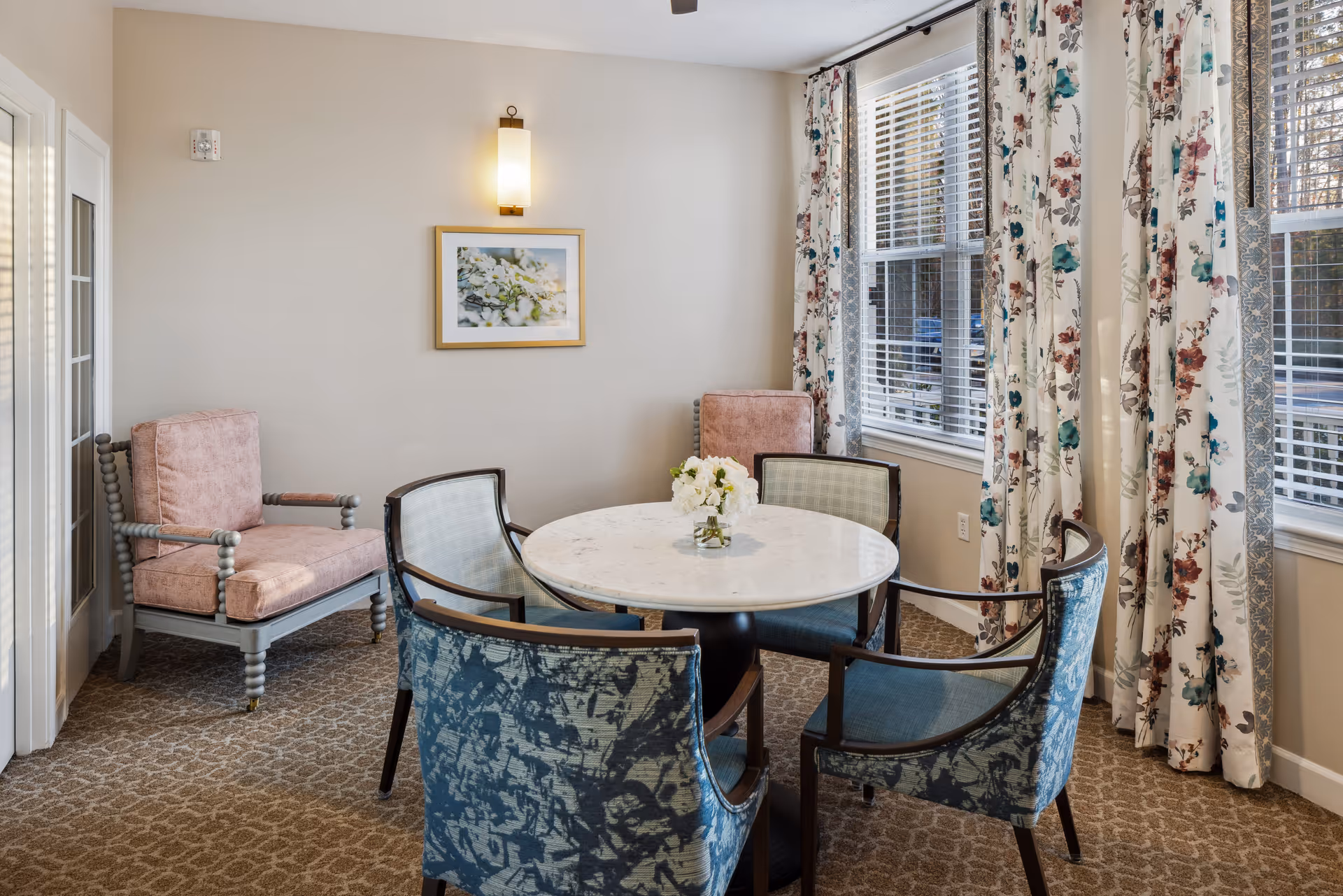 A cozy sitting area with a round marble table surrounded by four upholstered chairs with blue patterned fabric. Two pink cushioned armchairs are placed against the wall. The room has beige walls, a patterned carpet, floral curtains on the windows, and a framed picture with a wall sconce above it.