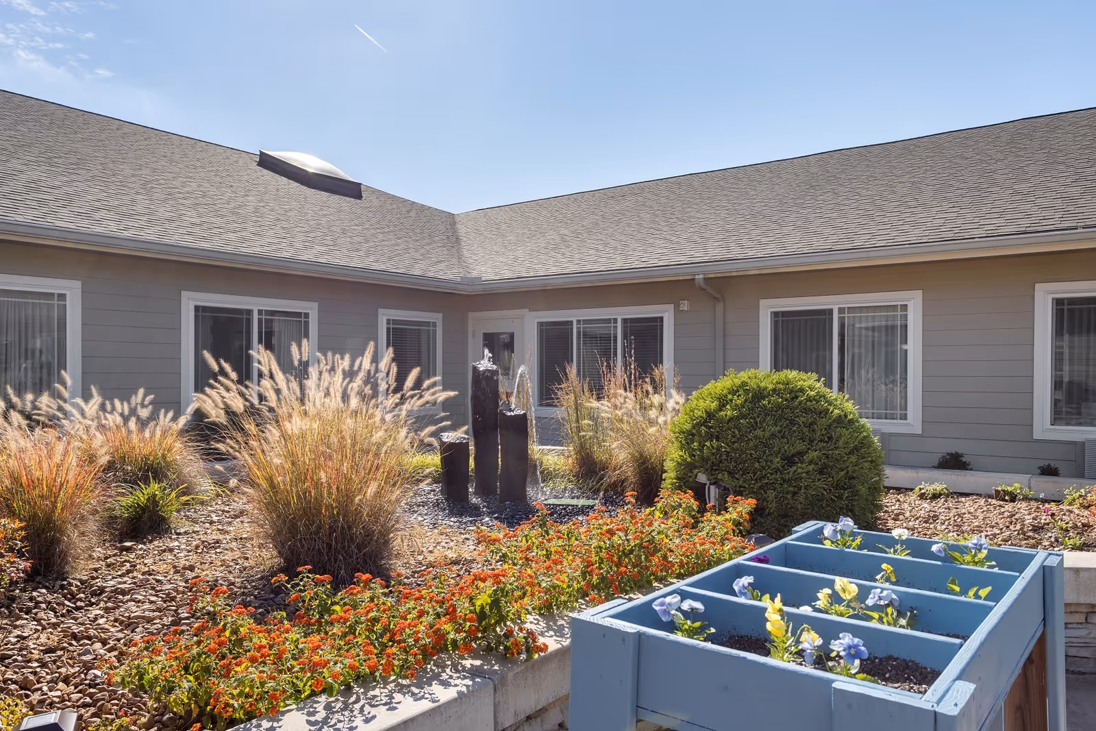 Outdoor garden area at Quail Ridge Assisted Living & Memory Care featuring ornamental grasses, colorful flowers, a small water fountain with three vertical stone pillars, and a blue planter box with blooming flowers, all set against the backdrop of a single-story building with multiple windows under a clear blue sky.