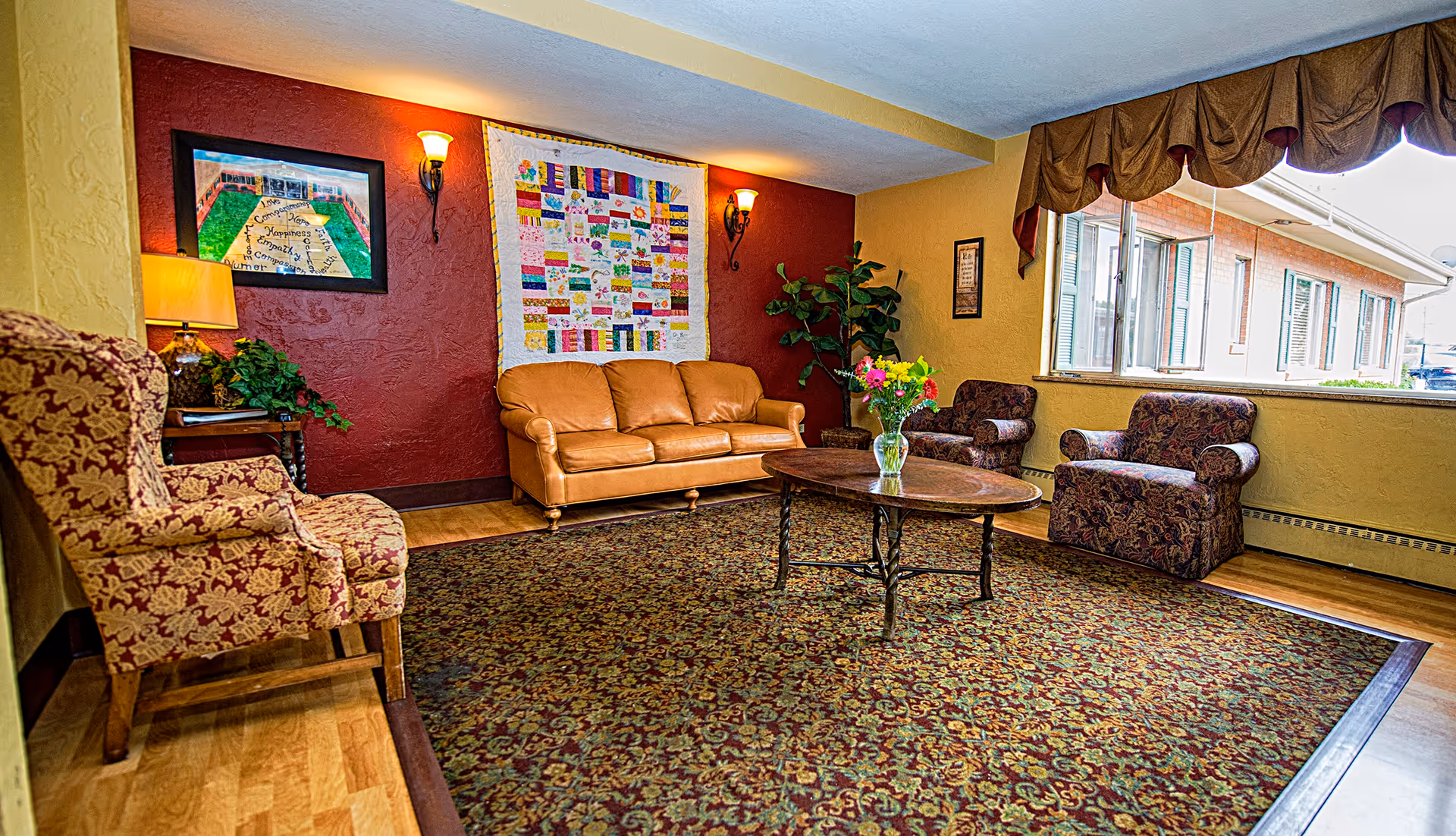 A cozy common lounge with a leather sofa, patterned armchairs, a coffee table, area rug, wall art and a large window.