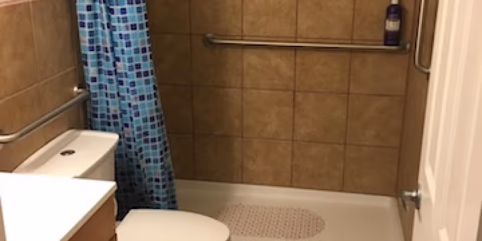 A bathroom with beige tiled walls, a white toilet, a white sink with a wooden cabinet, a bathtub with a blue patterned shower curtain, and metal grab bars installed on the walls for support. A bottle of soap or shampoo is placed on a shelf in the shower area.