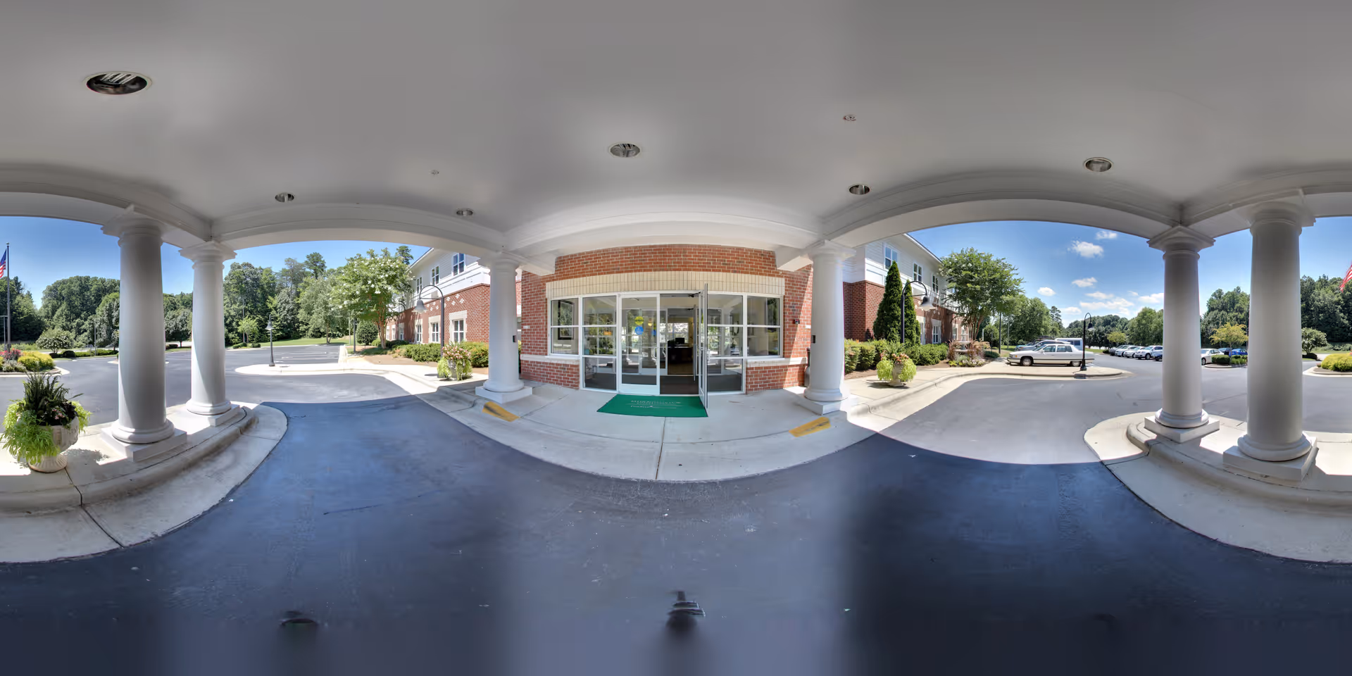 Entrance of a senior living facility with a covered driveway supported by large white columns. The building has a brick facade with glass doors and windows. There are potted plants near the columns and a parking lot with cars and greenery in the background under a clear blue sky.
