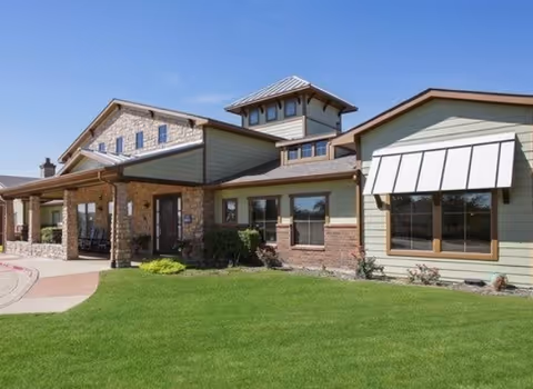 Exterior view of a single-story assisted living facility building with a combination of stone and siding walls, large windows, a covered entrance with pillars, and a well-maintained green lawn under a clear blue sky.