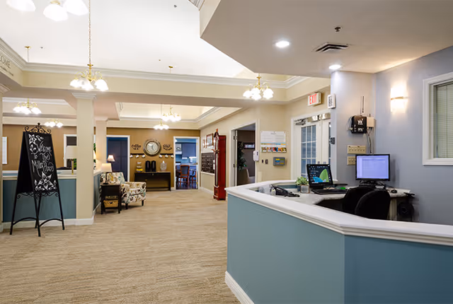 Reception desk and lobby area of a senior living facility with seating, chandeliers, and a hallway.