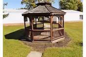 A wooden gazebo with a shingled roof situated on a grassy lawn area with a white building in the background under a clear sky.