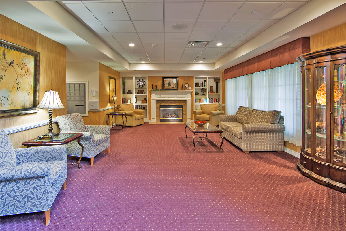 A spacious senior living facility lounge with patterned armchairs and a sofa arranged around a glass coffee table on a purple carpet. The room features a fireplace with built-in shelves on either side, decorated with books and ornaments. Large windows with sheer curtains allow natural light to fill the room, and a wooden display cabinet with glass doors is visible on the right.