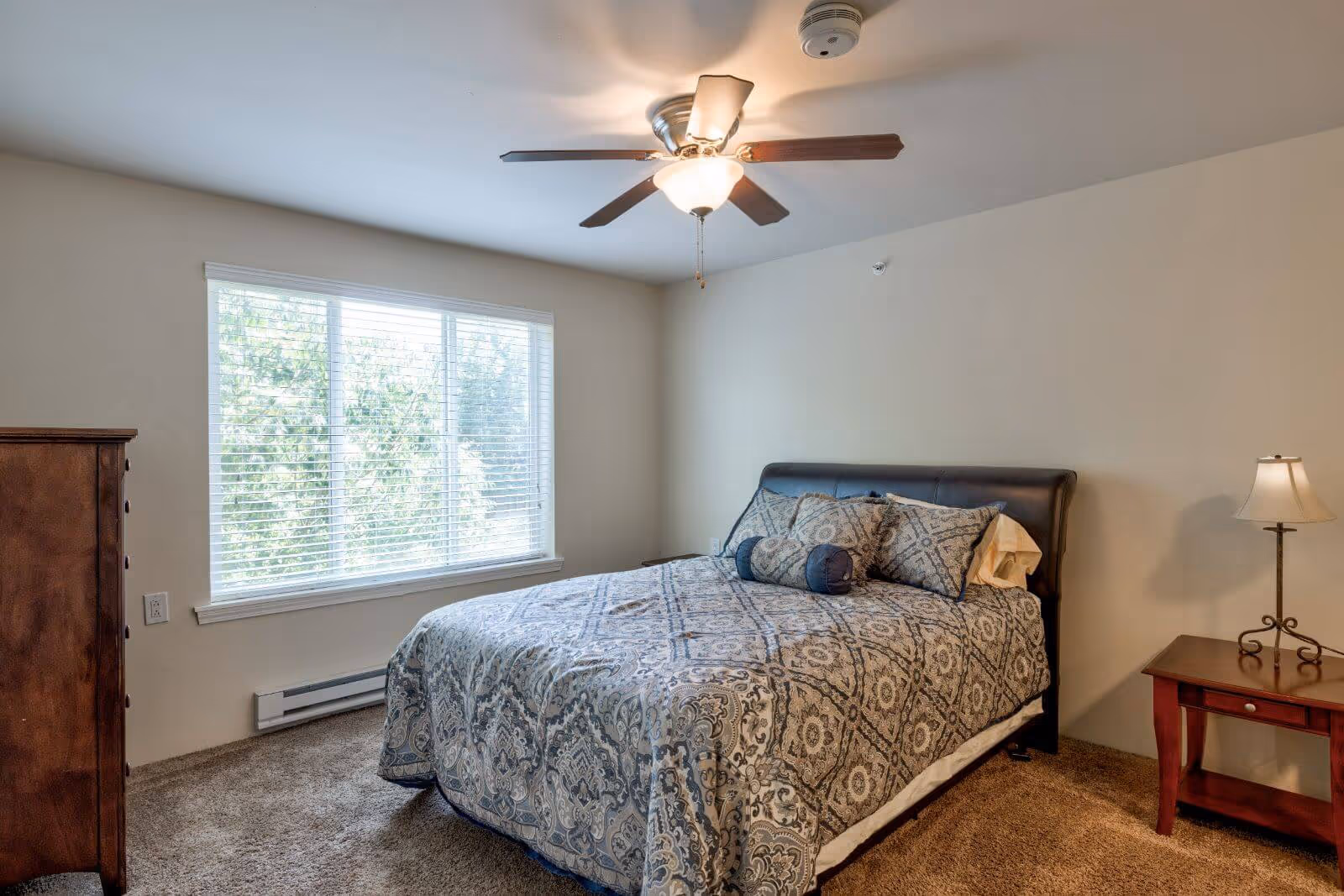 A bedroom with a large window covered by white blinds, a ceiling fan with light, a bed with patterned bedding and multiple pillows, a wooden nightstand with a lamp, and a wooden dresser on the left side.