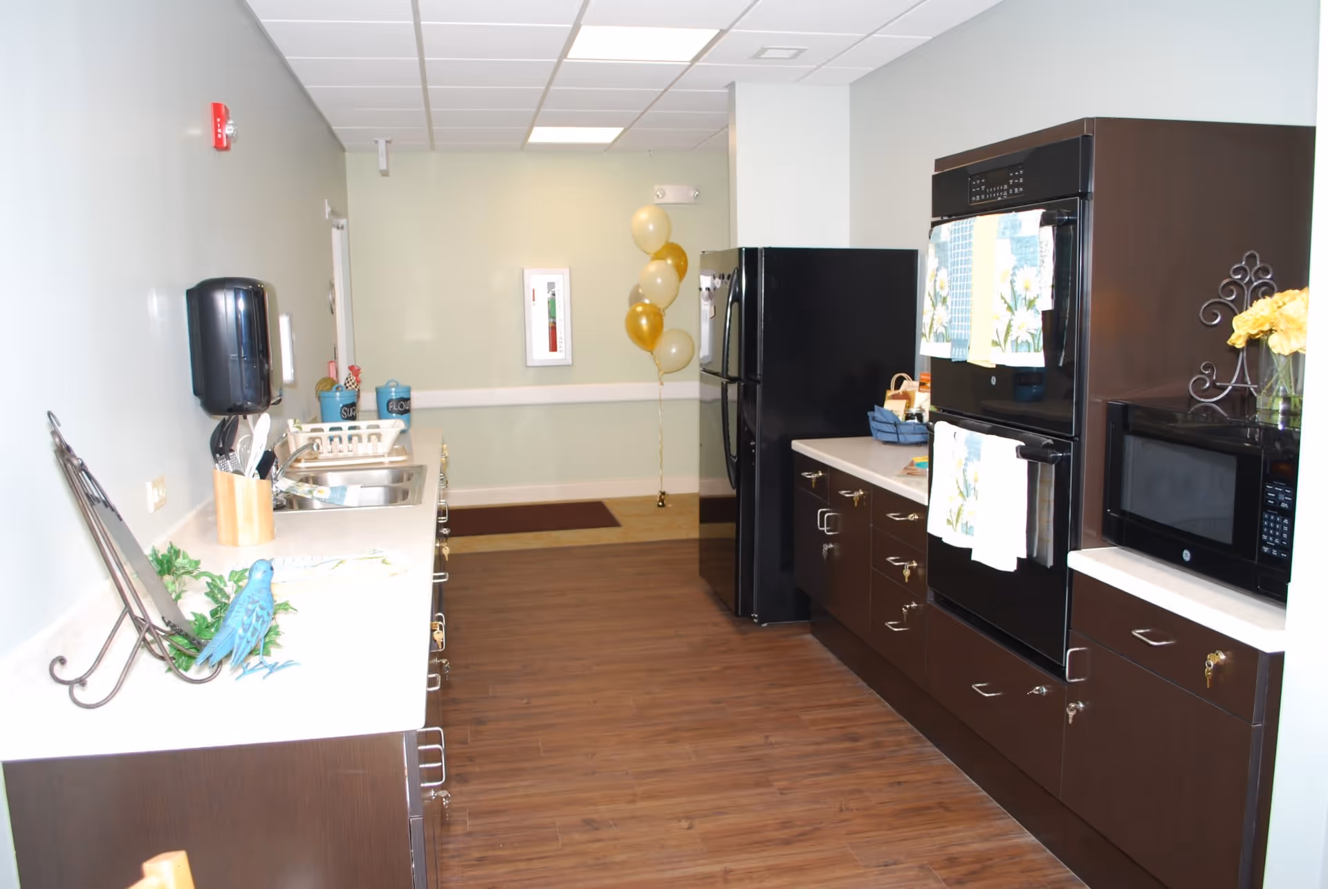 A modern kitchen area with dark wood cabinets and white countertops. The kitchen features a black refrigerator, a built-in double oven with floral-patterned towels hanging on the handles, a microwave, and a sink with utensils in a holder. There are decorative items including a blue bird figurine and yellow flowers. The floor is wood, and there are gold and white balloons in the background near a light green wall.