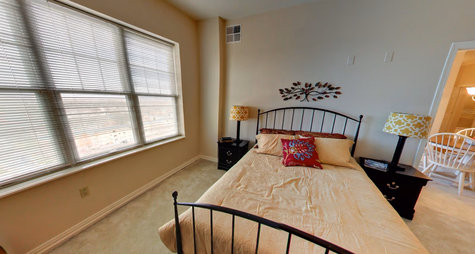 A bedroom with a large window covered by white blinds on the left side. The room features a bed with a black metal frame, beige bedding, and decorative pillows including a red one with a floral pattern. There are two black nightstands on either side of the bed, each with a lamp that has a yellow and white patterned lampshade. A decorative metal wall art piece shaped like a branch with leaves is mounted above the bed. In the background, a dining area with white chairs and a table is partially visible through an open doorway.