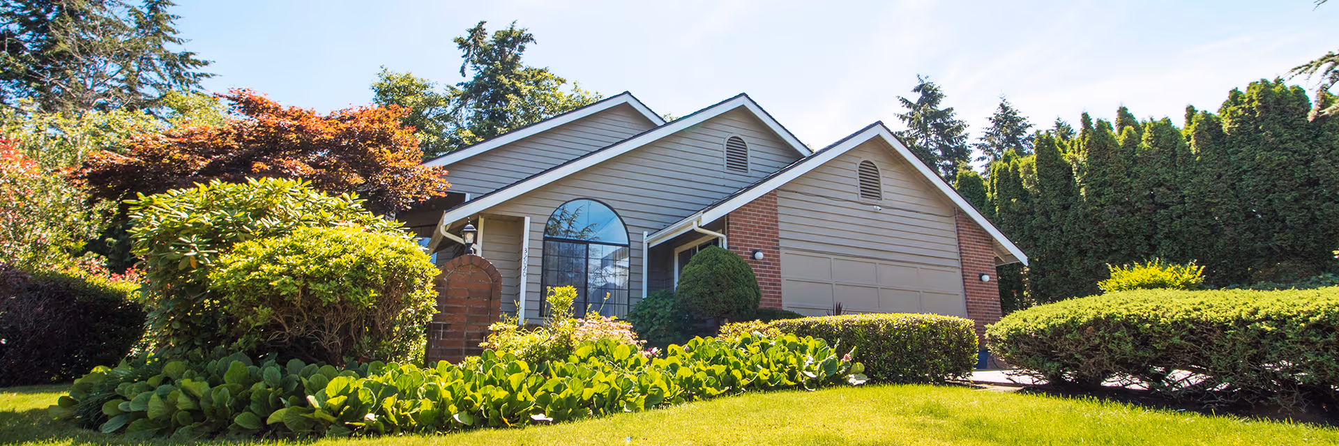 Exterior view of a single-story residential building with a garage, surrounded by well-maintained green bushes, trees, and a lawn under a clear blue sky.